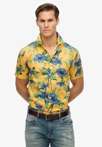 Camisa de manga corta amarilla con estampado floral de hibiscos azules y hojas verdes. Presenta un cierre de botones y un cuello clásico, combinada con unos vaqueros azules y un cinturón marrón.