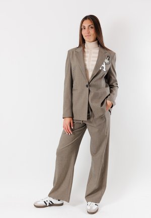 Completo grigio a quadri con giacca su misura caratterizzata da una toppa e pantaloni a gamba larga. Il dolcevita adornato aggiunge texture. Sneakers completano il look.