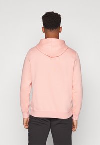 Sweat à capuche rose clair avec des manches longues, des poignets et un ourlet côtelés. Texture douce. Porté avec un pantalon gris foncé, vu de dos.