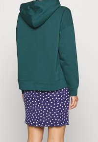 Mörkgrön hoodie med huva, avslappnad passform och ribbad kant, i kombination med en lila kjol med vit fjärilstryck och scalloped kant.