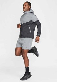 Nike lättviktsjacka i grått och svart med huva, tillsammans med grå shorts. Funktioner inkluderar dragkedjestängning och reflekterande detaljer.