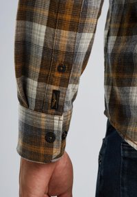 Braun- und graues kariertes Shirt mit Ärmel und Knopfmanschetten, das eine gewebte Textur aufweist und ein Etikett mit einem Logo an der Manschette trägt.