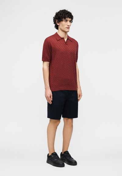 Άνδρας που στέκεται στραμμένος ελαφρώς προς τα δεξιά, φοράει κόκκινη πόλο μπλούζα με κοντά μανίκια και υφή, μαύρα σορτς μέχρι το γόνατο και μαύρα αθλητικά παπούτσια σε απλό φόντο.
