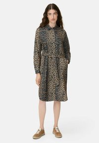 Frau steht mit einem langärmligen Kleid im Leopardenmuster, einem Gürtel, beigen Sneakers und ihrer linken Hand in der Tasche.