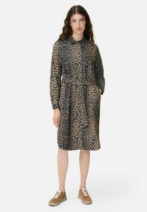 Kvinde står iført en langærmet leopardmønstret kjole med bælte, beige sneakers, og hendes venstre hånd i lommen.