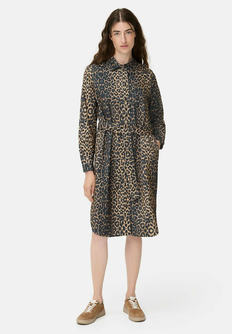Frau steht mit einem langärmligen Kleid im Leopardenmuster, einem Gürtel, beigen Sneakers und ihrer linken Hand in der Tasche.