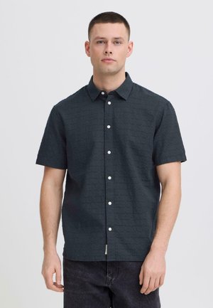 Jeune homme portant une chemise à manches courtes boutonnée texturée foncée et un pantalon noir, debout devant un fond clair uni.