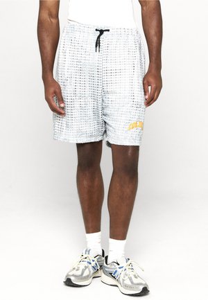 Uomo che indossa pantaloncini bianchi con texture, coulisse nera e dettaglio giallo, calzini bianchi e scarpe sportive argento e blu.