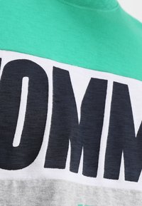 Camisa corta de manga corta en verde y blanco con un gran texto en azul marino. La tela parece suave, con bloques de color horizontal en gris que contrastan.