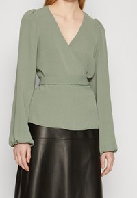 Blouse verte à manches bouffantes longues et taille ceinturée, associée à une jupe en cuir noire. Le tissu présente une finition texturée subtile.