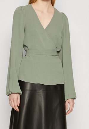 Groene wikkelblouse met lange pofmouwen en een ceintuur in de taille, gecombineerd met een zwarte leren rok. De stof heeft een subtiele textuur.