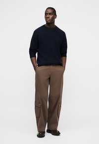 Άνδρας που στέκεται με τα χέρια στις τσέπες, φοράει μπλε σκούρο πουλόβερ, καφέ παντελόνια καρό και σκοτεινά παπούτσια σε απλό λευκό φόντο.