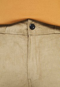 Beiga kordfladderbyxor med ett strukturerat vertikalt randmönster, med en svart knappstängning och en synlig, slät orange skjorta ovanför.