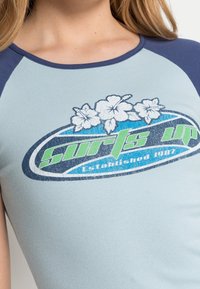 Světle modrá košile s krátkým rukávem se modrozeleným grafickým logem s květinami a textem "Curls Up Established 1987" tučně.
