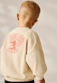 Dziecko z krótkimi blond włosami noszące beżowy sweter z czerwoną grafiką, na której na plecach widnieje napis "Red Hot Chili Peppers By The Way".