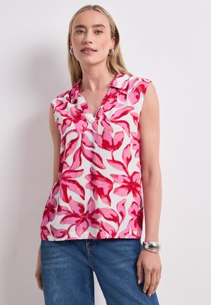 Femme portant un chemisier blanc sans manches avec imprimé floral rose et rouge, jean bleu, bracelet argenté et boucles d'oreilles créoles, debout contre un mur blanc.