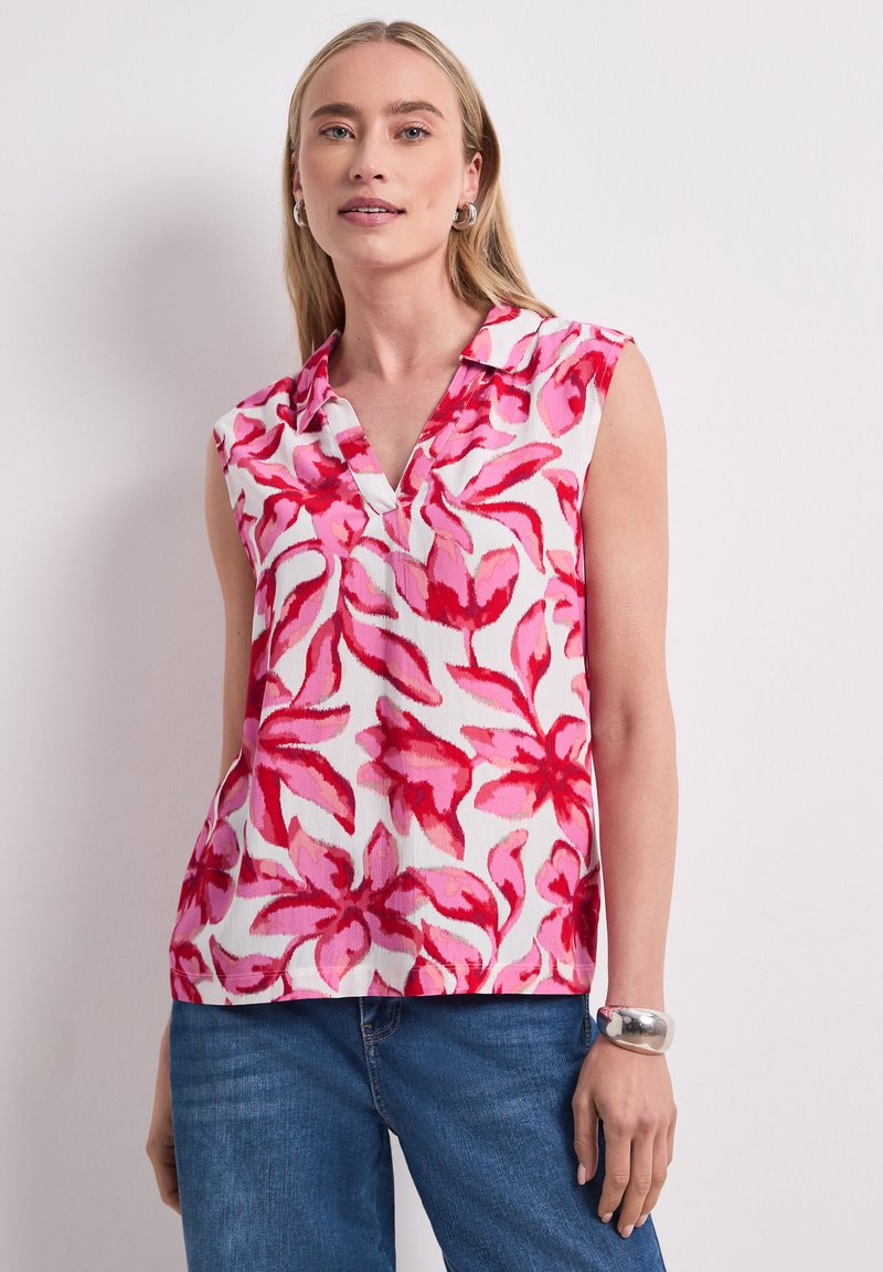 Mujer con blusa blanca sin mangas con estampado floral rosa y rojo, vaqueros azules, pulsera plateada y pendientes de aro, de pie contra una pared blanca.