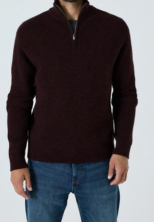 Homme portant un pull zippé bordeaux foncé et un jean bleu, debout devant un fond clair uni.