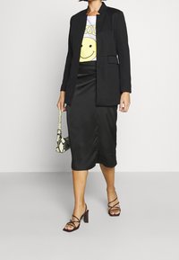 Svart kavaj över en vit t-shirt med ett gult leende ansikte, kombinerad med en svart satinkjol och sandaler med remmar och klack. Handväska i ormskinn ingår.