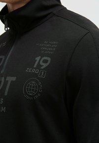 Schwarze Zip-Jacke aus strukturiertem Material mit Drucken in verschiedenen Farbnuancen und auffälligen Textdesigns, darunter "19 ZERO 1" und ein Globus-Logo.
