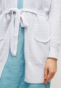 Cardigan lavorato a maglia di un grigio chiaro con un motivo perforato, cintura da annodare e due tasche frontali, indossato sopra un vestito blu.