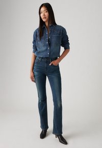 Denim marškiniai su dviem priekinėmis kišenėmis, mygtukų užsegimu ir ilgomis rankovėmis. Tamsiai mėlynos džinsai su siauru siluetu ir lengvu išsiplėtimu apačioje. Juodi aukštakulniai batai.