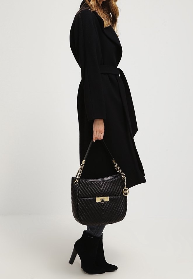 Femme en manteau noir et bottines à talons tenant un sac à main noir matelassé avec chaîne et fermoir dorés.