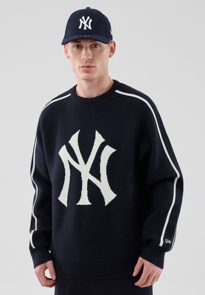 Νέος άνδρας που φοράει σκούφο New York Yankees σκούρου μπλε χρώματος και ταιριαστή μπλε μπλούζα με μεγάλο λευκό λογότυπο NY και λευκές ρίγες στα μανίκια.