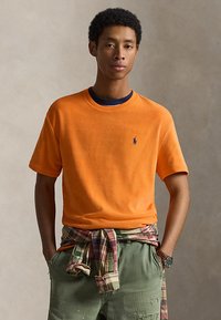 Νεαρός άνδρας που φορά πορτοκαλί κοντομάνικο πουκάμισο με ναυτικό λογότυπο, καρό πουκάμισο δεμένο στη μέση, χακί παντελόνι και ρολόι χειρός, στέκεται με τα χέρια στις τσέπες.