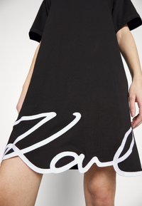 Svart bomullst-shirtklänning med korta ärmar och rundad nederkant. Stort vitt broderat "Karl"-detalj vid nederkanten. Slät yta.