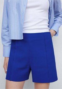 Pantalones cortos azules a medida con una textura suave, que presentan una cintura alta, paneles laterales y líneas limpias, combinados con una camisa a rayas azul clara.