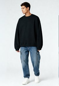 Fekete, oversized pulóver bordás mandzsettával és szegéllyel, világoskék cargo farmerrel, oldalzsebekkel és fehér sportcipőkkel párosítva.