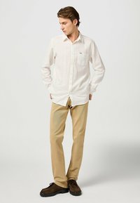 Witte overhemd met lange mouwen, knoopsluiting aan de voorkant en één borstzak, gecombineerd met tan kleurige broek en donkerbruine schoenen.