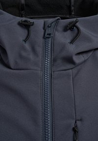 Close-up van de rits van een marineblauwe jas met zwarte verstelbare trekkoorden op de stoffen capuchon en een klein ritszakje aan de rechterkant.