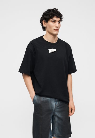 Νέος με σγουρά μαλλιά, φοράει μια μαύρη ευρύχωρη μπλούζα με λευκή ετικέτα και σκούρα τζιν σορτς, στέκεται με το ένα χέρι στην τσέπη.