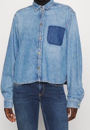 Personne portant une chemise en jean bleu clair à boutons avec une poche poitrine en jean bleu foncé sur un jean bleu foncé et un col roulé noir.