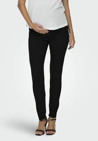 Jean skinny de maternité noir, tissu extensible, coupe slim, texture lisse, doté d'une taille mi-haute, associé à des sandales à talons hauts à lanières noires.