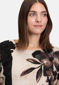 Jeune femme aux cheveux bruns lisses portant un pull en tricot beige avec de grands motifs floraux sombres et de petites perles décoratives.