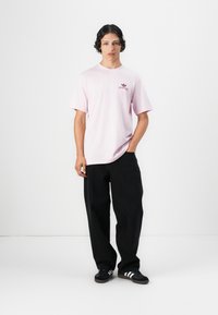 Rosa Baumwoll-T-Shirt mit kurzen Ärmeln und Rundhalsausschnitt, das ein kleines rotes Logo auf der Brust trägt, kombiniert mit schwarzen weit geschnittenen Hosen und schwarzen Sneakers.
