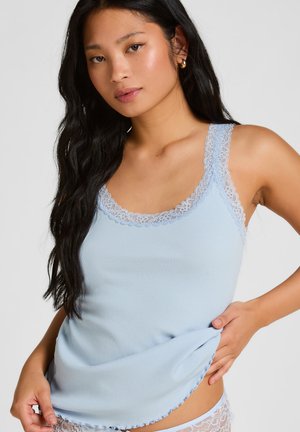 Jonge vrouw met lang donker haar, gekleed in een lichtblauw tanktop met ribbelstructuur en kanten randje, houdt het zoom van haar top iets omhoog aan haar zijde.