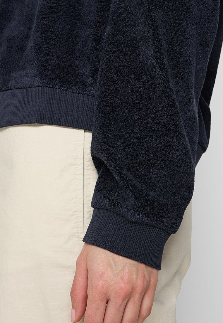 Dunkelblauer Fleecepullover mit weicher Textur, gerippten Bündchen und einem lockeren Schnitt. Getragen über hellen Hosen.