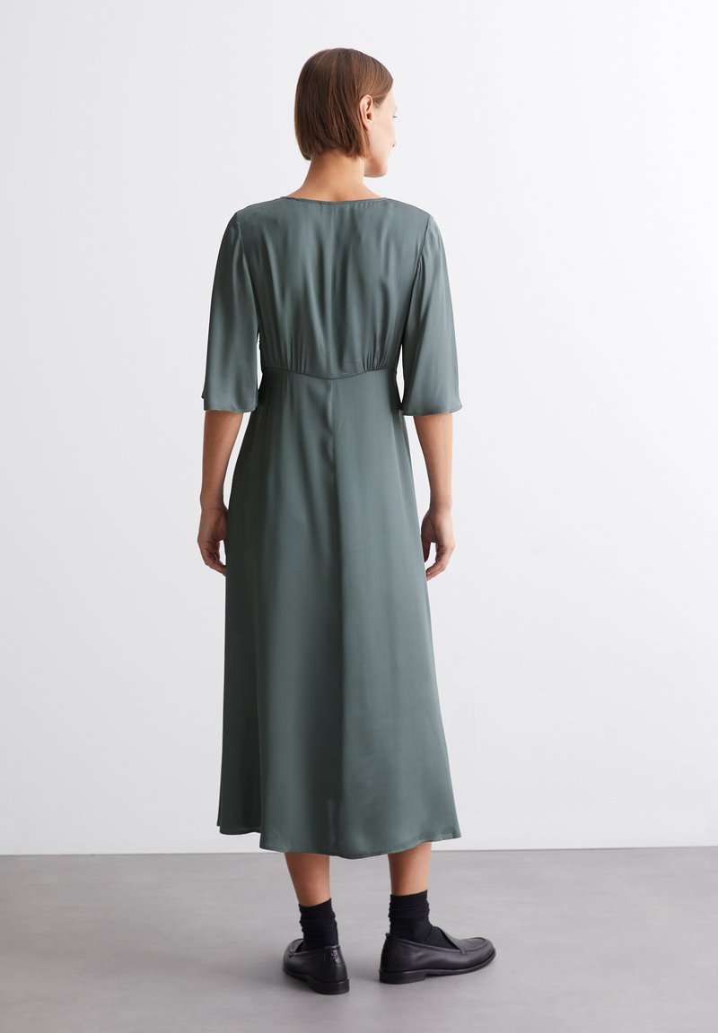 Langes, olivengrünes Kleid mit einer weichen, glatten Textur. Hat eine taillierte Taille und dreiviertellange Ärmel. Getragen mit schwarzen Schuhen.