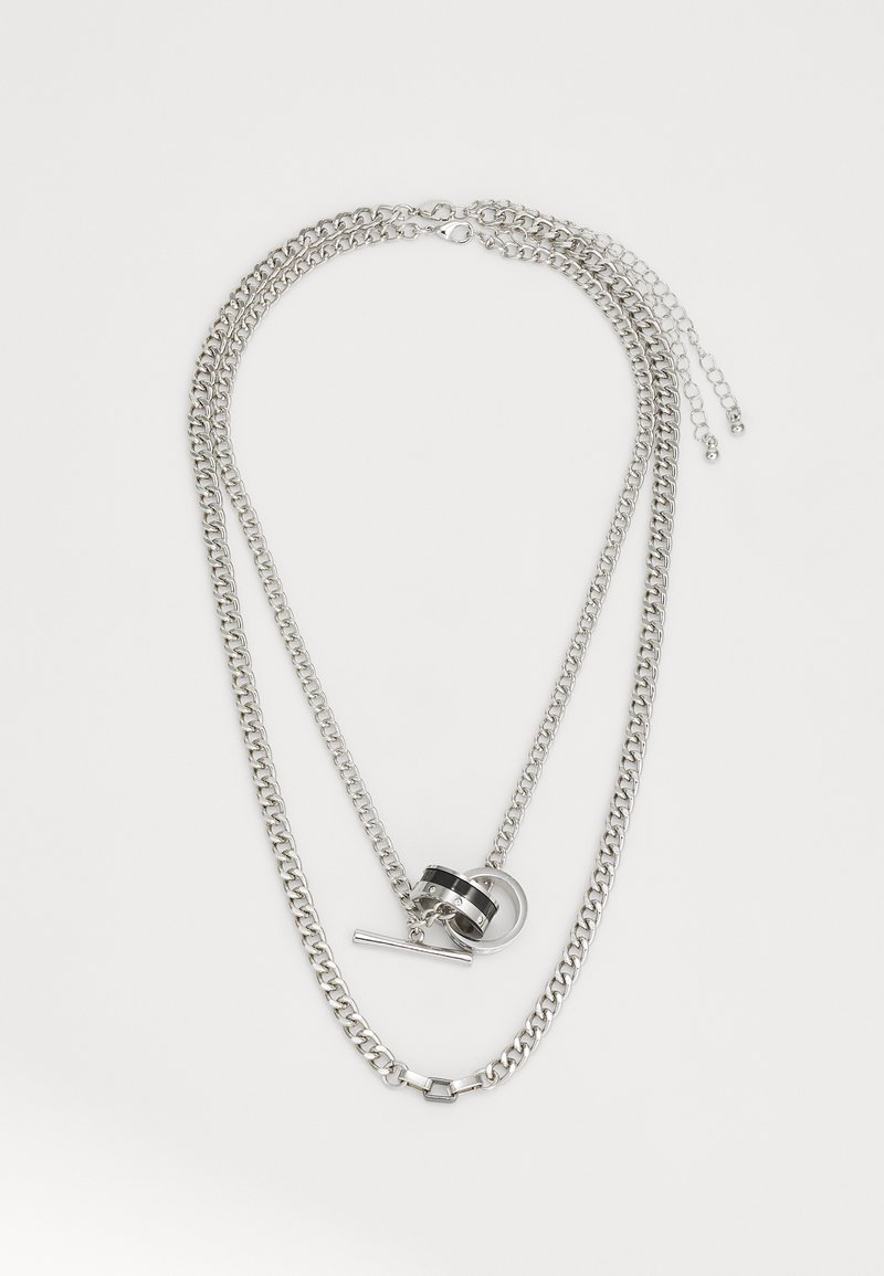 Collier en chaîne multi-couches argenté avec des maillons de tailles variées, mettant en avant un pendentif noir et argenté ainsi qu'un détail de fermoir à bascule.