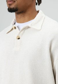 Cream-colored knit pullover with a textured finish, featuring a collared design and two light brown buttons at the neckline.