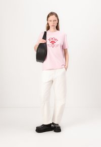 Roza majica s rdečim logotipom "JOOP! JEANS", v kombinaciji z belimi širokimi hlačami in črnimi platform čevlji. Model drži črno torbo čez ramo.