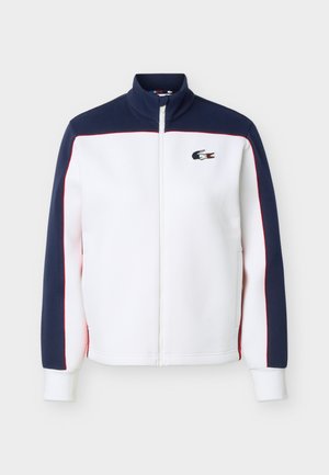 Bílá bunda na zip s tmavě modrými rameny a rukávy, červeným lemováním, vysokým límcem a logem krokodýla Lacoste na levé straně hrudníku.