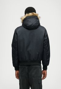 Μαύρο μπουφάν bomber με αποσπώμενη κουκούλα επενδεδυμένη με γούνα, ελαστικούς καρπούς και στρίφωμα. Λείος υφασμάτινος φινίρισμα με εφαρμοστή σιλουέτα.