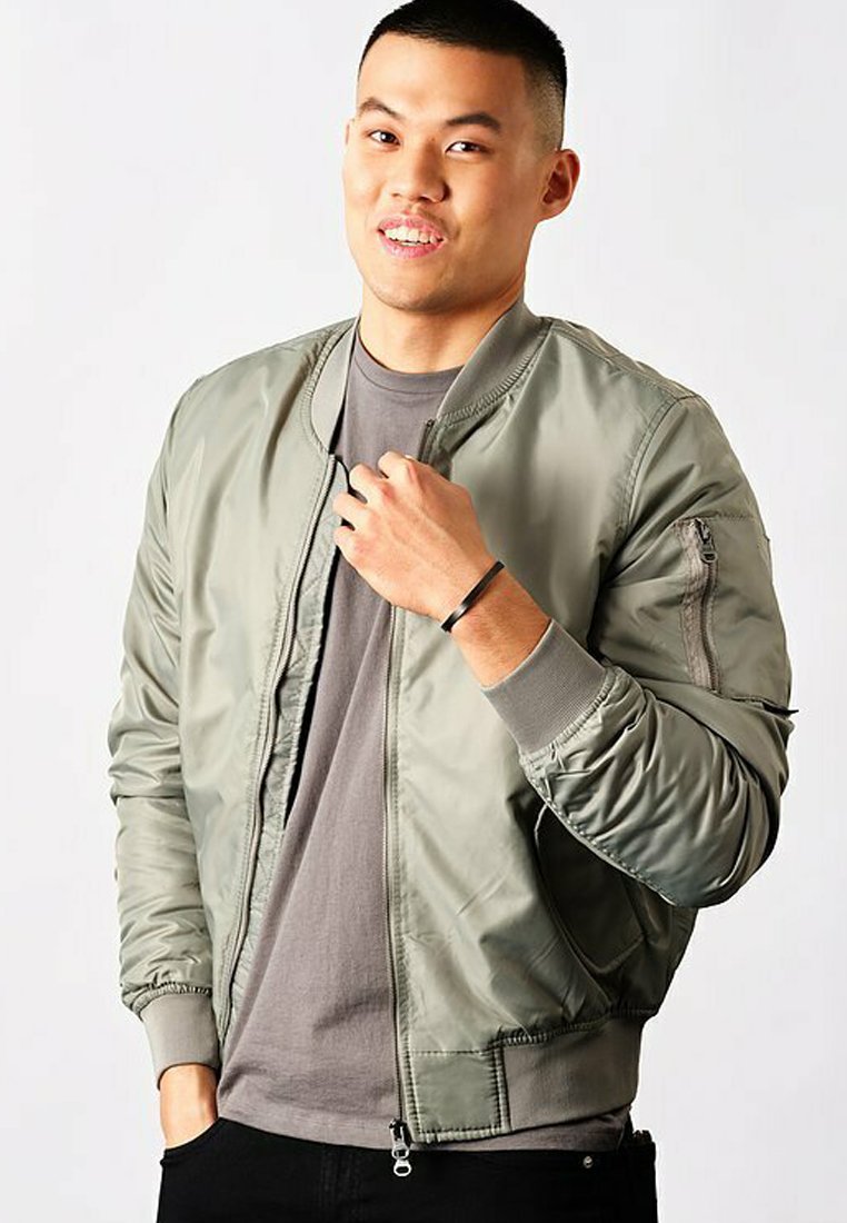 Veste bomber vert olive avec une texture lisse et brillante, fermeture éclair à l'avant, poignets côtelés et une poche sur la manche gauche, portée sur une chemise grise.