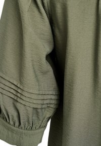 Olivgrönt tyg med en mjuk struktur, med en veckad design på ärmen och en rynkad mudd vid handleden. Lätt och avslappnad passform.