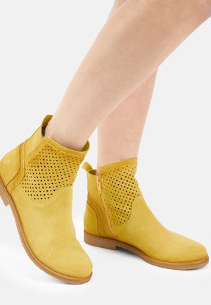 Gule ruskind ankelstøvler med perforerede oversider, lynlåslukning på siden og lave brune gummisåler, båret på bare fødder.